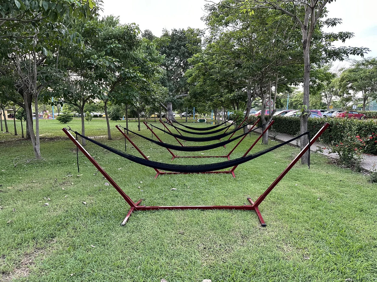 Secuencia de hamacas oscuras en soportes individuales, alineadas sobre el césped entre árboles.