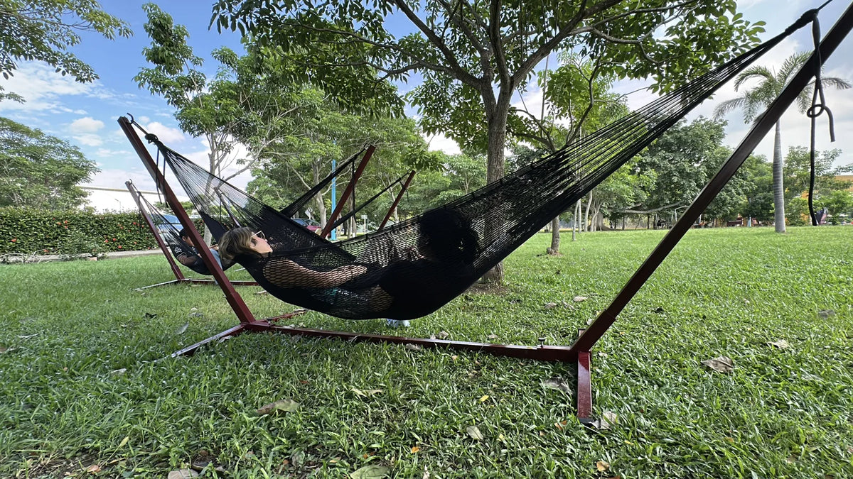 Una persona descansa en una hamaca oscura sobre el césped del campus, bajo los árboles.