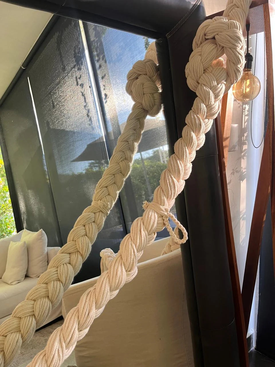Trenzas claras suspendidas en diagonal frente a una ventana, junto a sillones blancos.