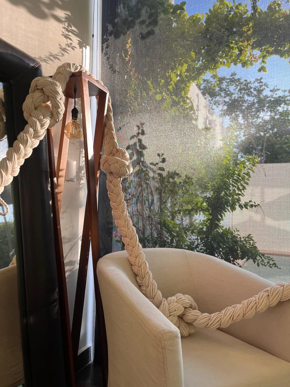 Remate de la composición de trenzas junto a un sillón y una ventana con vegetación al exterior.