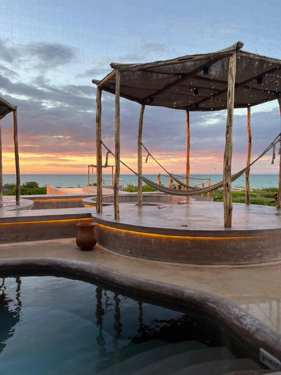 Hamacas bajo un pabellón de madera junto a una piscina circular al atardecer.