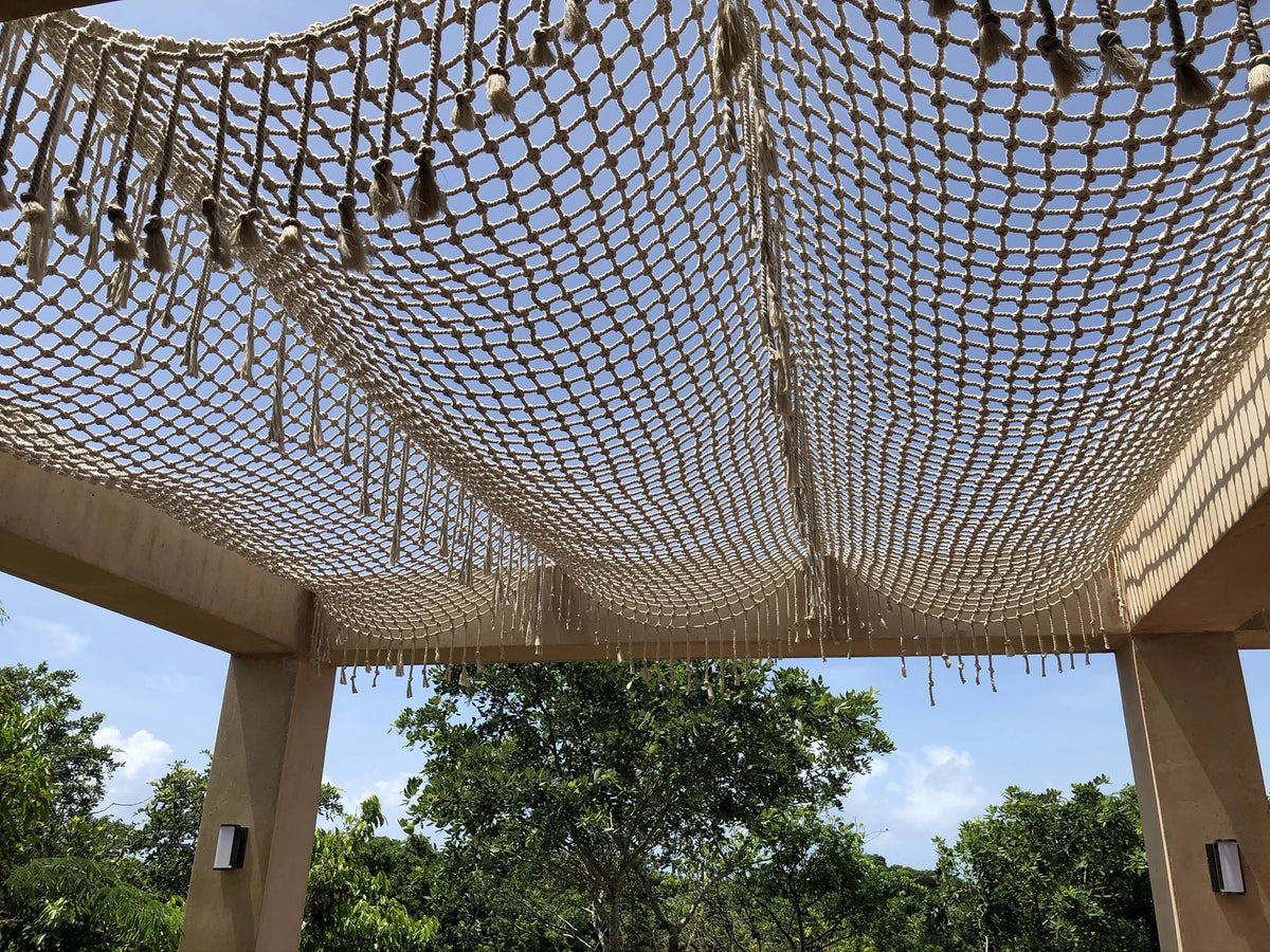 Detalle de la red tejida y sus flecos sobre los pilares de una pérgola, con cielo azul y árboles detrás.