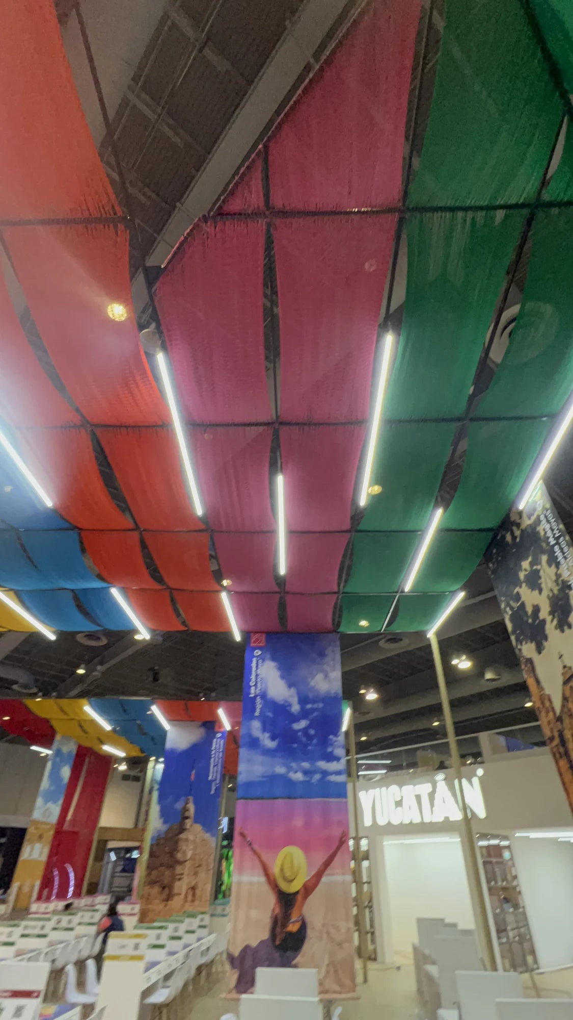 Vista desde el interior del stand de Yucatán, con franjas de tejido verde, rosa, naranja y azul suspendidas sobre las áreas de encuentro.