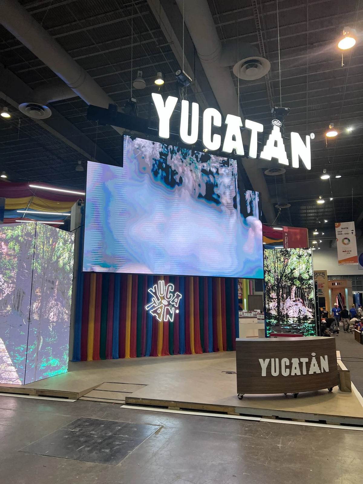 Frente del stand de Yucatán, con cortinas multicolor bajo una pantalla y un letrero luminoso.